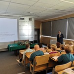 Lek. Marcin Malka podczas swojej prezentacji. Na pierwszym planie plecy uczestników. Na drugim planie wykładowca, stół prezydialny, rzutnik. Wykładowca stoi pod oknami zasłoniętymi roletami, w ręku trzyma bezprzewodowy mikrofon.