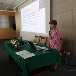 Prof. dr hab. n. med. Waleria Hryniewicz przy stole prezydialnym, na którym jest laptop, kompozycja z kwiatów, woda, szklanki oraz dokumenty. Na drugim planie rzutnik, na którym wyświetlana jest prezentacja.