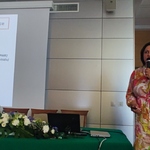 Dr n. med. Alicja Kuch przy stole prezydialnym, na którym jest laptop, kompozycja z kwiatów, woda, szklanki oraz dokumenty. Na drugim planie rzutnik, na którym wyświetlana jest prezentacja.